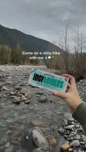 A hand holding an incredibles chocolate bar beside a mountain creek, captioned: come on a rainy hike with me
