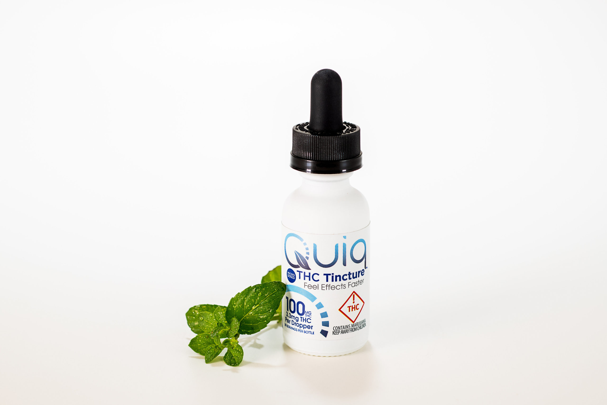 Quiq fast-acting THC-infused tincture, made by Medically Correct, is resting on a white surface with a small mint leaf to the side of the bottle.