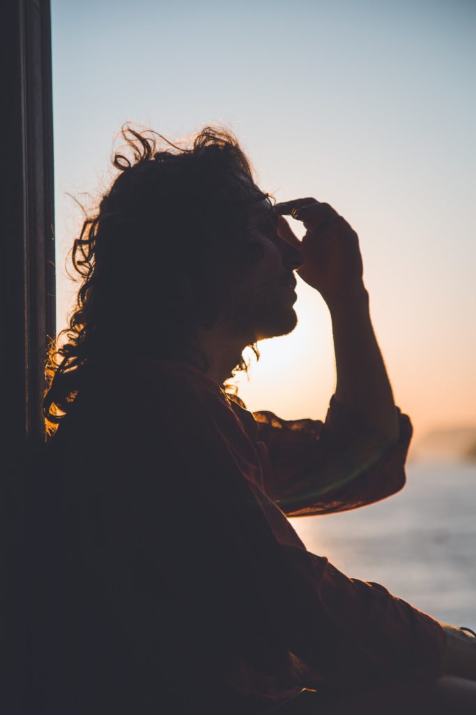 Shadowy person with curly hair and a long sleeved blouse sitting outside at sunset holding their hand on their forehead to display stress before smoking cannabis to relieve chronic pain